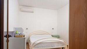 Ein kleines Zimmer mit einem Einzelbett mit Metallgestell, einer weißen Wand, einem kleinen Kühlschrank und einer grünen Mülltonne.
