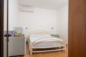 A small, single-bed room with a metal frame bed, a white wall, a small refrigerator, and a green trash bin.