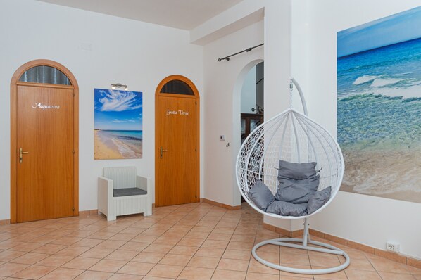 A room with two wooden doors labeled 'Acqua Viva' and 'Grotta Verde,' a white chair, a hanging egg chair, and a beach mural.