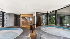 Ein moderner Poolbereich im Innenbereich mit zwei Whirlpools, einer Treppe und großen Fenstern, die einen Blick auf die umliegenden Gebäude bieten.