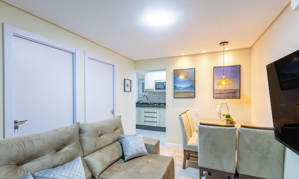 A living room with a sofa, a dining area with a table and chairs, a kitchen in the background, and a TV mounted on the wall. - Apto 2Qtos centro BC a 160m da praia SRL002 (Balneário Camboriú)