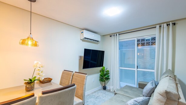 A modern living room with a sofa, a dining table with chairs, a TV mounted on the wall, and a large window with curtains. - Apto 2Qtos centro BC a 160m da praia SRL002 (Balneário Cambori ú)