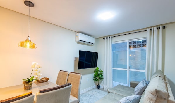 A modern living room with a sofa, a dining table with chairs, a TV mounted on the wall, and a large window with curtains.