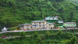 A multi-story hotel with a green roof, located on a hillside surrounded by lush greenery. - DALLAS THE RETREAT RESORT (Mussoorie)