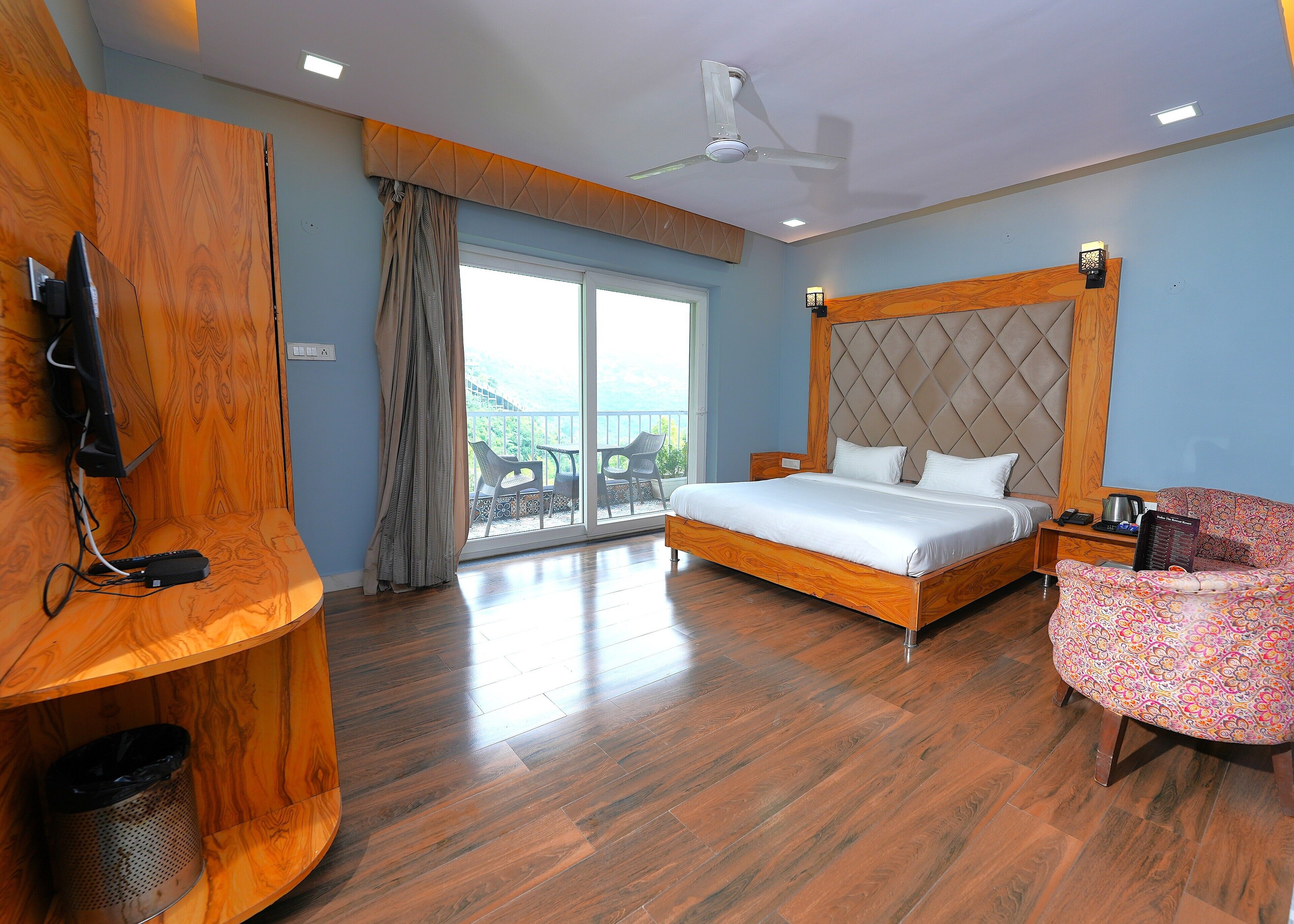 A hotel room with a large bed, a TV mounted on a wooden panel, a balcony with chairs, and a ceiling fan.