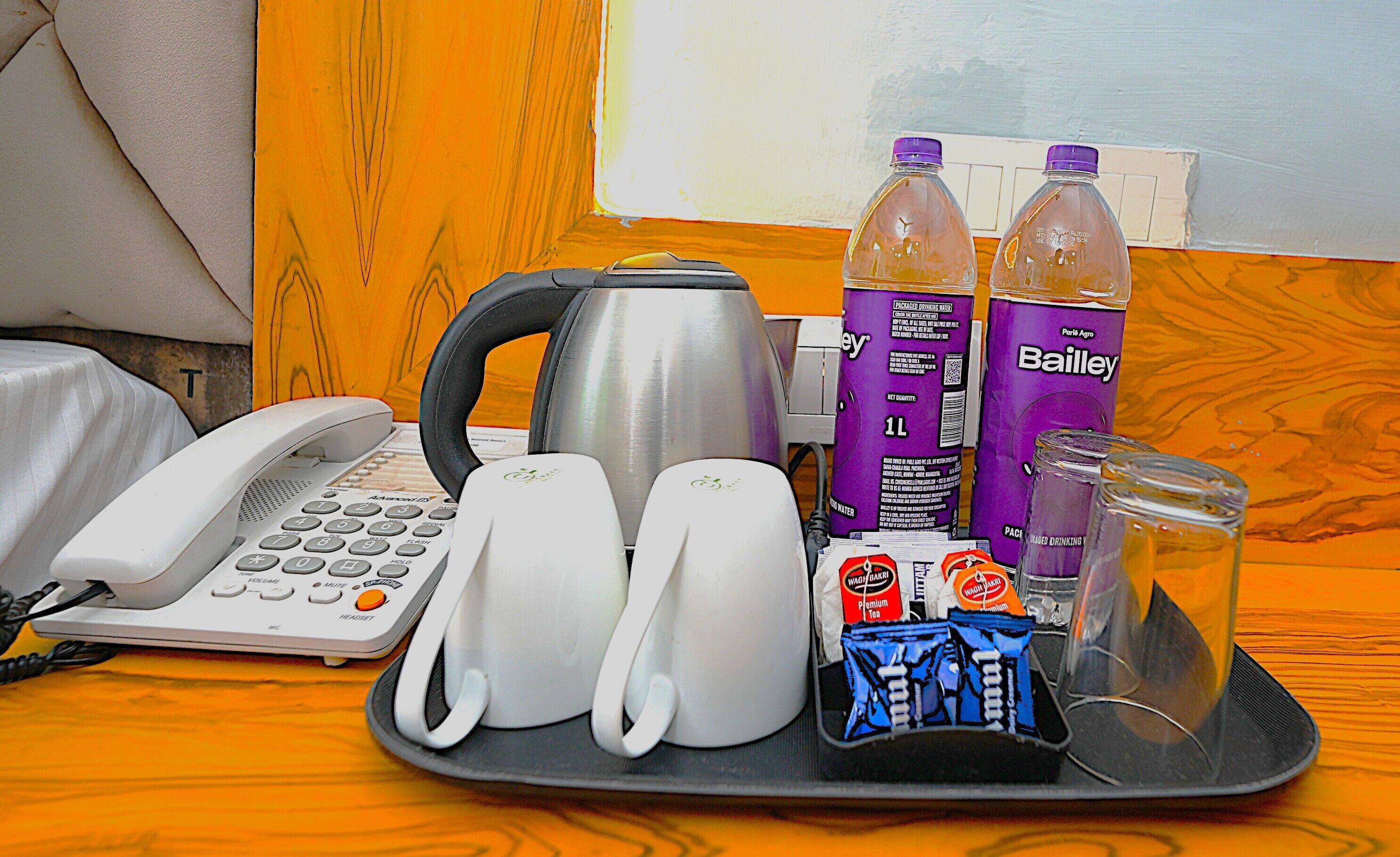 A hotel room with a telephone, a kettle, two Bailey water bottles, a tray with snacks, and a glass.