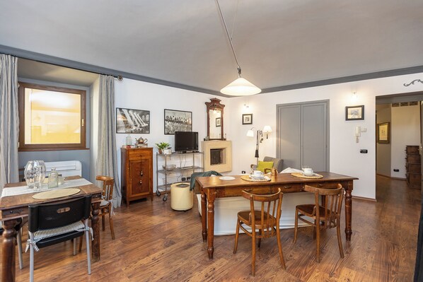 A dining area with a wooden table, chairs, and a chandelier. There is a television on a stand, a cabinet, and a window with curtains. - Rental in Rome Pantheon Apartment (Rome)