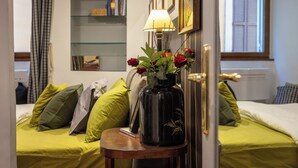 A room with a bed, a bedside table with a lamp and a vase of flowers, a bookshelf, and a window with shutters. - Rental in Rome Pantheon Apartment (Rome)