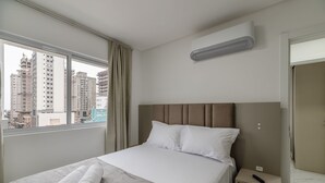 A hotel room with a bed, a bedside table, a remote control, and a view of tall buildings through a window. - 2BR apt w/ BBQ 700m From the Beach Sir0501 (Itapema)