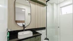 A modern bathroom with a wooden panel wall, a large oval mirror, a black countertop, and a glass shower enclosure. - 2BR apt w/ BBQ 700m From the Beach Sir0501 (Itapema)