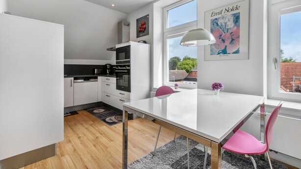 A modern kitchen with white cabinets, a black countertop, and a white dining table with a pink chair. - Apartment '3' Villa Elysium (Heide)