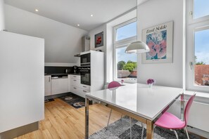 Cocina moderna con armarios blancos, encimera negra y mesa de comedor blanca con una silla rosa.