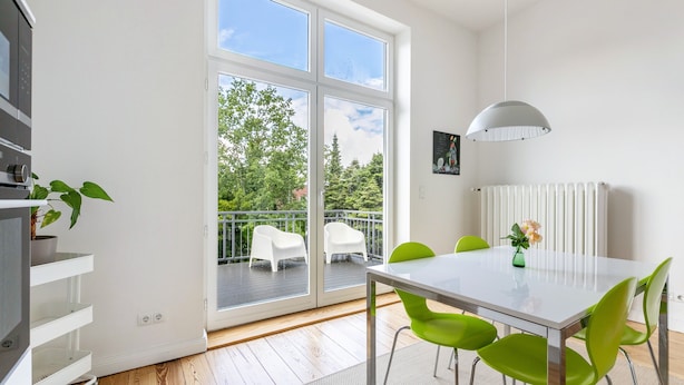 A modern kitchen with a built-in oven, a dining area with a white table and green chairs, and a balcony with white chairs and a view of greenery.