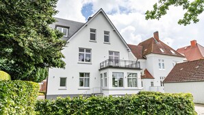 A white two-story house with a balcony, surrounded by greenery.