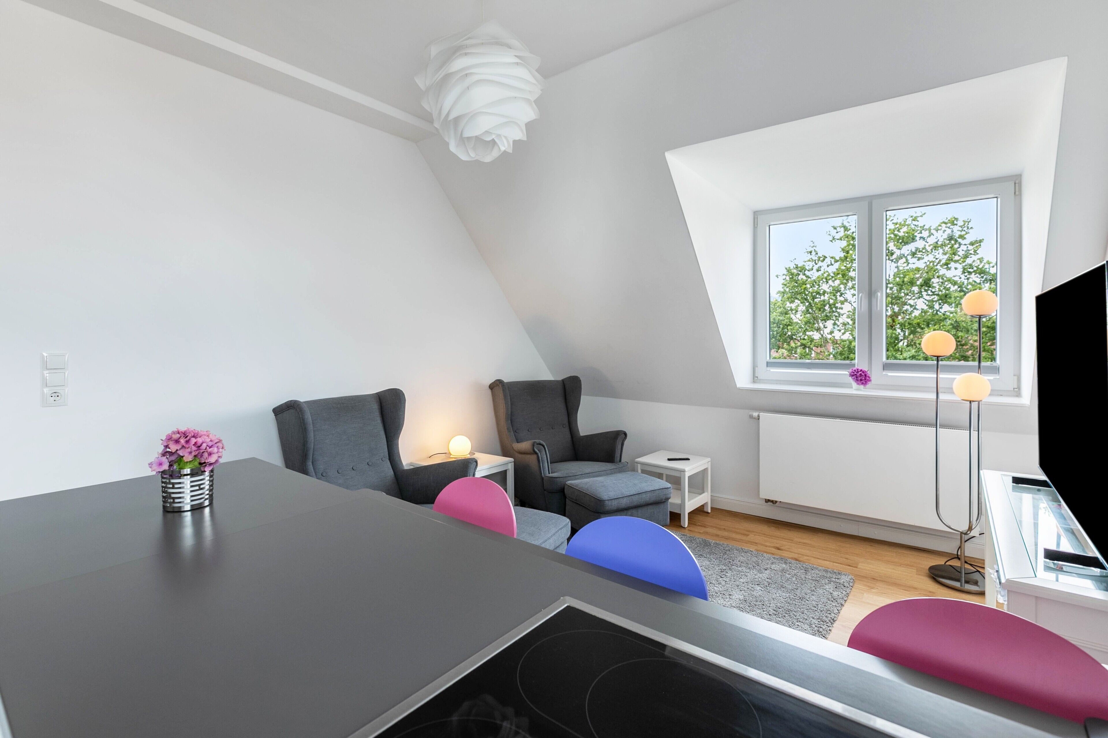 Camera da letto in stile loft moderna con soffitto a falde, un tavolo da pranzo, un piccolo tavolino bianco, una lampada da terra e una finestra con vista sugli alberi.