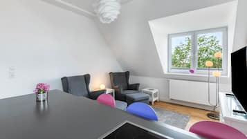 Camera da letto in stile loft moderna con soffitto a falde, un tavolo da pranzo, un piccolo tavolino bianco, una lampada da terra e una finestra con vista sugli alberi.