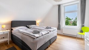 A neatly made bed with pillows and blankets, a bedside table with a lamp, a window with curtains, and a bright green chair. - Apartment '4' with Wi-Fi (Heide)