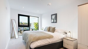 A modern bedroom with a large bed, a bedside table with a lamp, and a view of the outdoors through a large window. - Axis Apartment (Leeds)