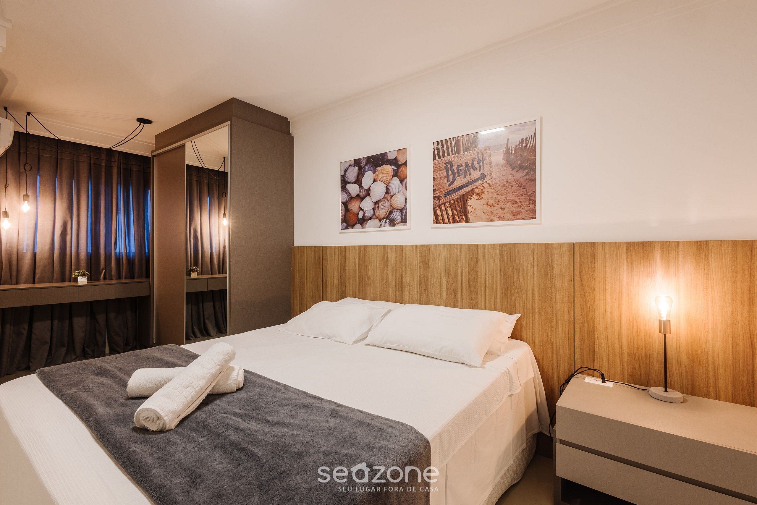 Uma cama bem arrumada com um cobertor cinza e toalhas dobradas, uma mesa de cabeceira com abajur, e duas imagens emolduradas na parede.