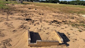 Un campo arenoso con una estructura de madera, cielo despejado y edificios a lo lejos.