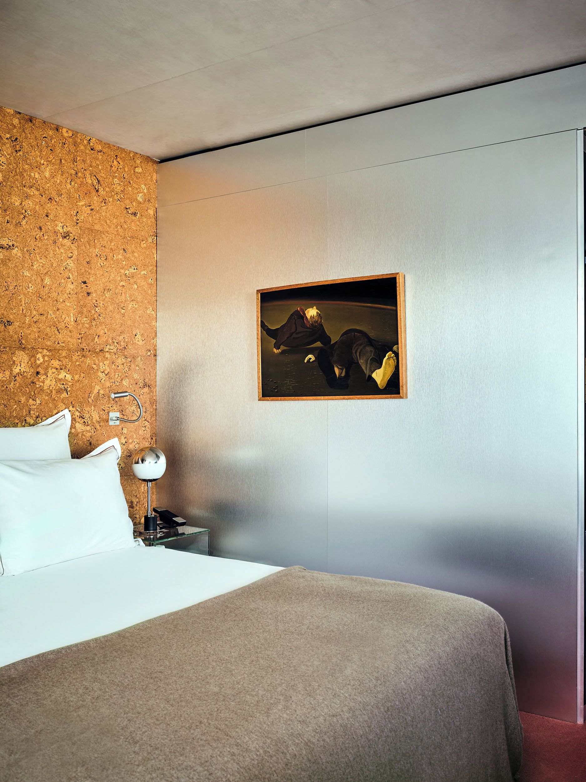 A hotel room with a bed, a nightstand with a lamp, a painting on the wall, and a corkboard.