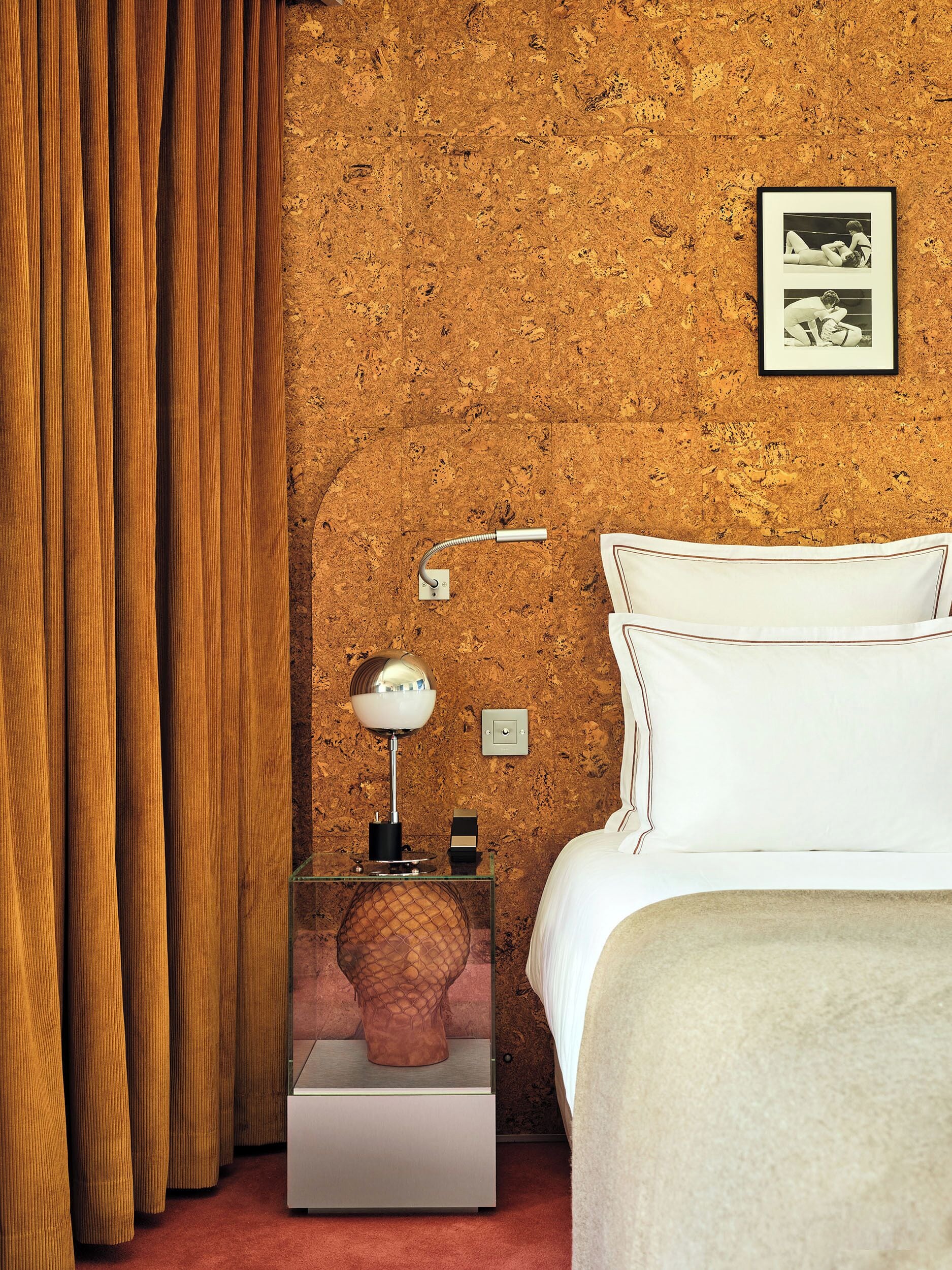 A bedroom with a cork wall, a bed with white bedding, a nightstand with a lamp, and a framed picture.