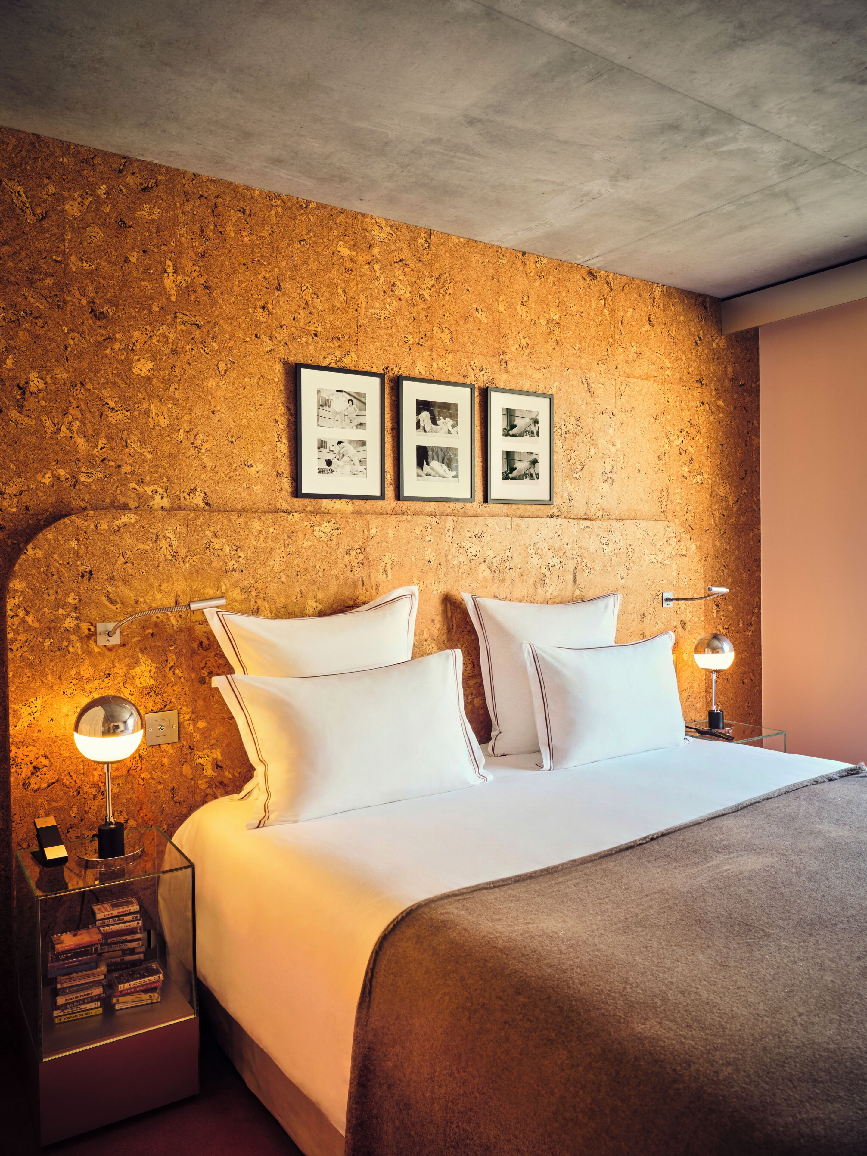 A hotel room with a bed, two bedside lamps, a wall-mounted shelf with books, and three framed pictures on the wall.