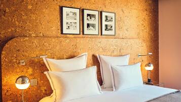 A hotel room with a bed, two bedside lamps, a wall-mounted shelf with books, and three framed pictures on the wall.