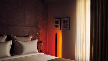Une chambre d’hôtel moderne avec un grand lit, une table de chevet, une lampe et une vue sur l’extérieur à travers des rideaux légers.