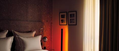 A modern hotel room with a large bed, a nightstand, a lamp, and a view of the outdoors through sheer curtains.