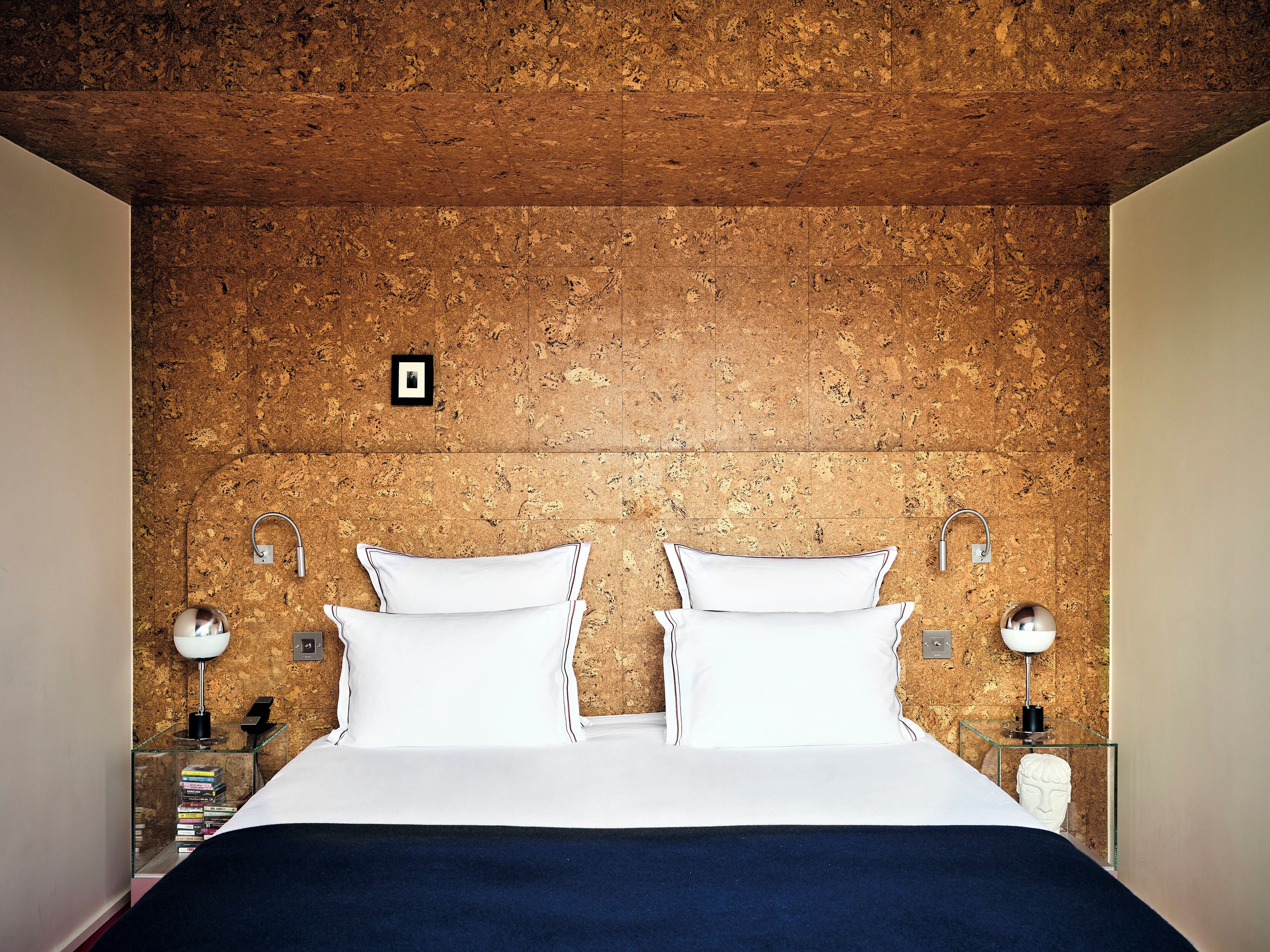 A bedroom with a cork wall, two white pillows, a bed, and two bedside lamps.