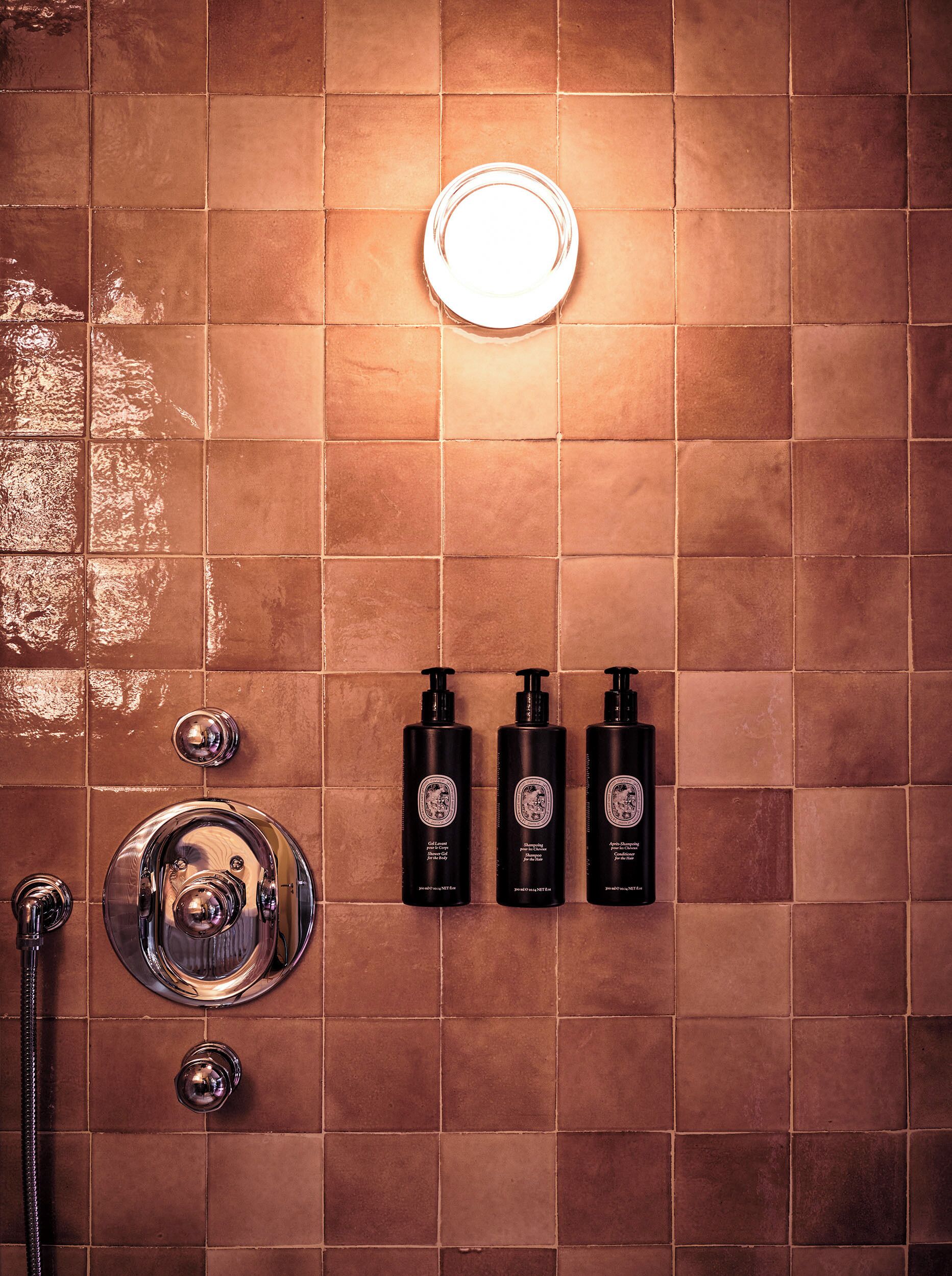 A bathroom with a tiled wall, a round mirror, a handheld showerhead, and three black bottles of shampoo.