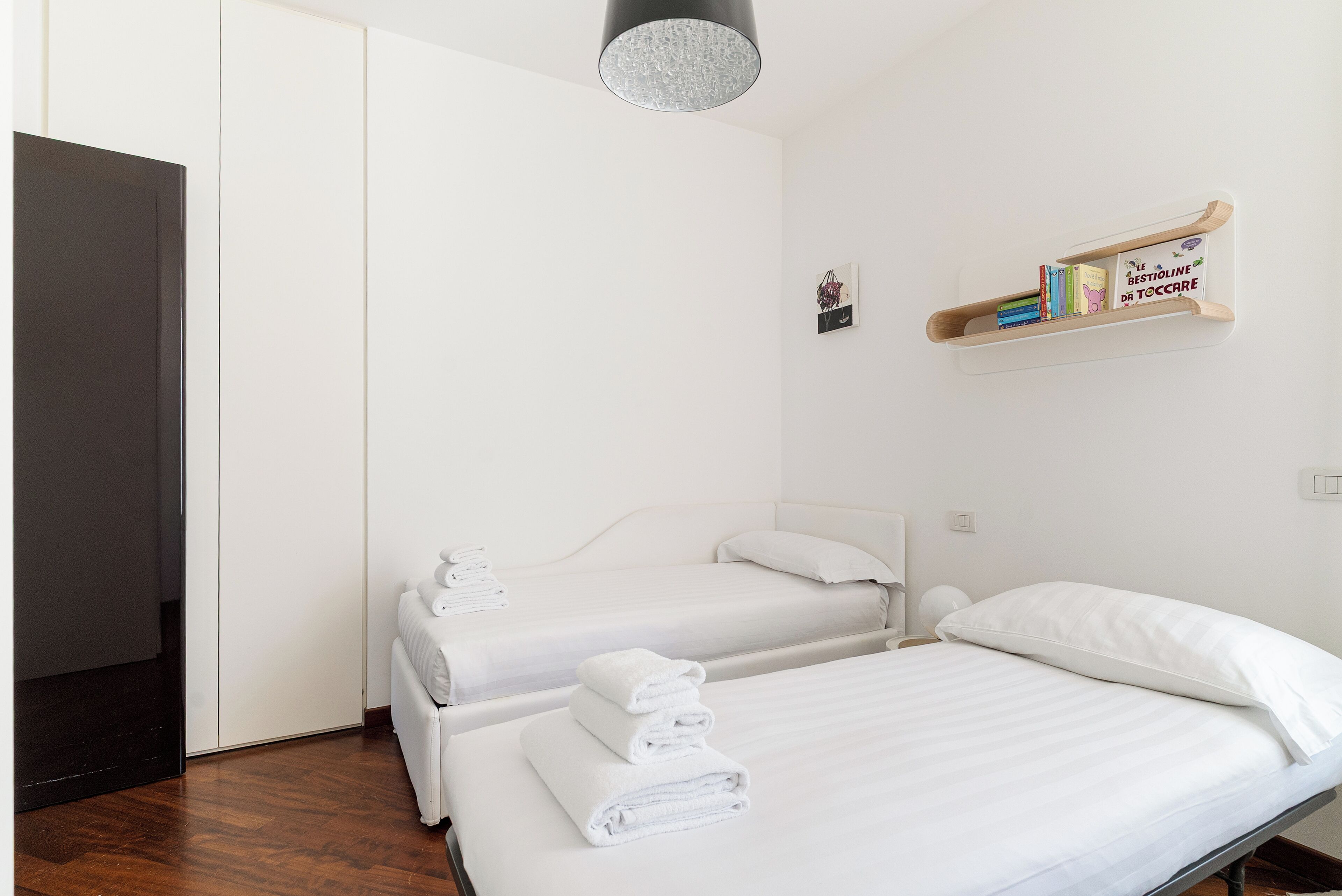 A hotel room with two beds, white linens, a bookshelf with books, and a hanging light fixture.