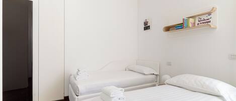 A hotel room with two beds, white linens, a bookshelf with books, and a hanging light fixture.