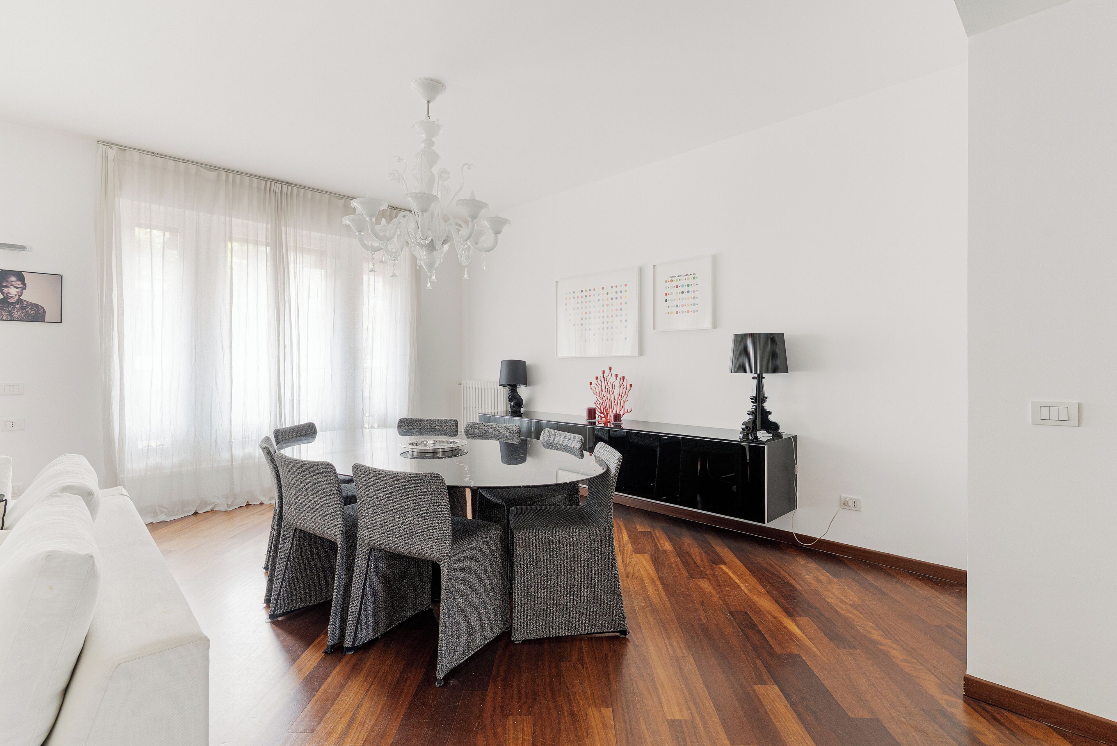 A modern, well-lit living room with a round dining table, grey upholstered chairs, a white sofa, a chandelier, and a black entertainment unit.