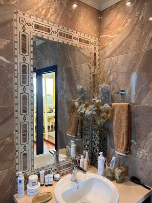 Un baño con pared de mármol, un espejo grande, un lavabo y artículos de aseo.