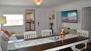 Un comedor con una mesa de madera, sillas blancas, un mantel corrido floral y un televisor en la pared.