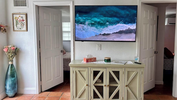Una sala de estar con una televisión montada en un mueble, un florero con flores y un cuadro en la pared.