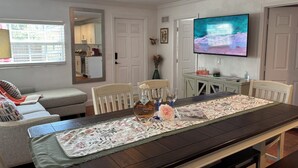 Un comedor con una mesa larga dispuesta para seis personas, un televisor montado en la pared y una cocina visible a través de una puerta abierta.