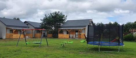 Un espacio al aire libre con césped, un columpio, un trampolín y un edificio de madera con techo inclinado.