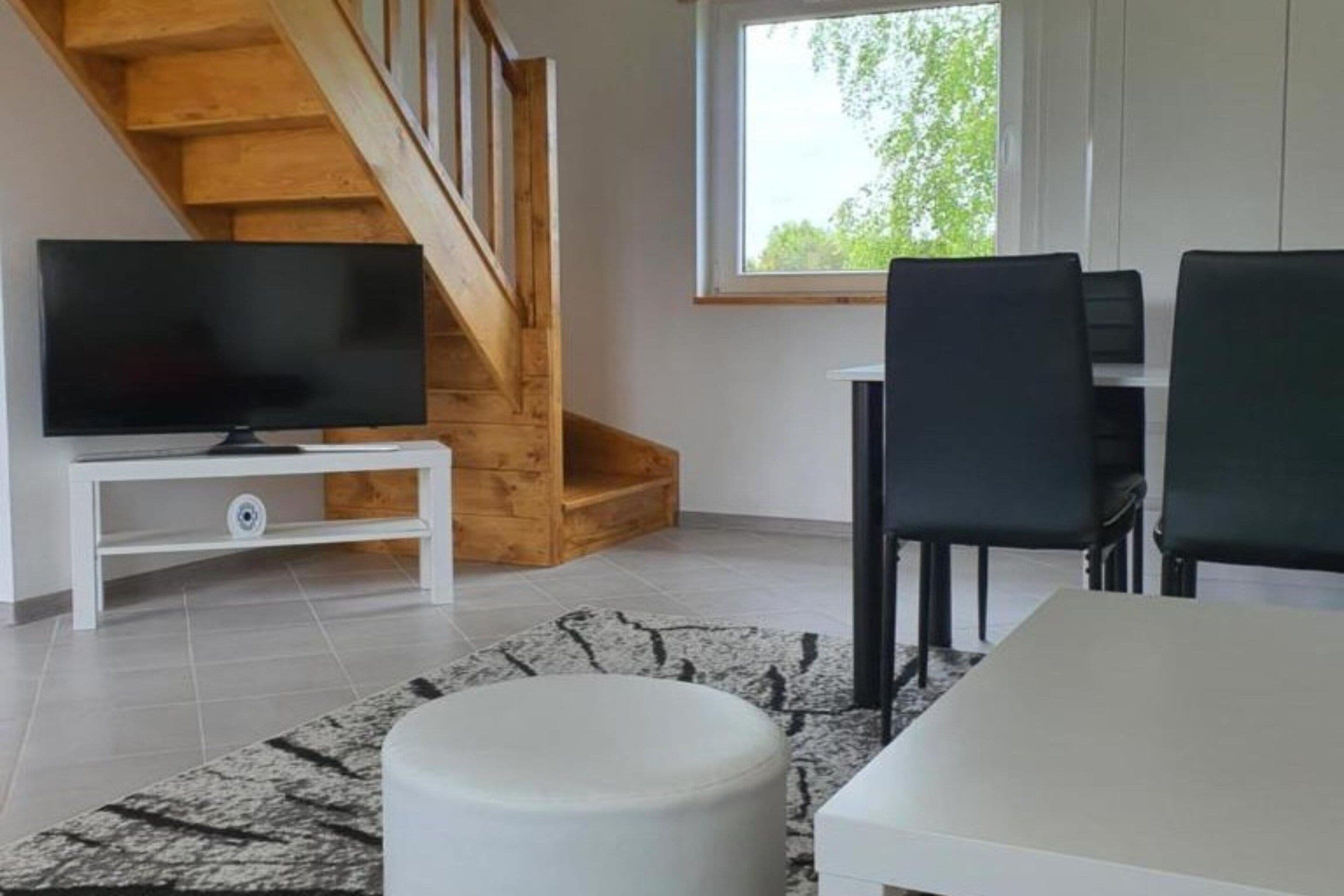 A modern living room with a TV on a white stand, a dining table with chairs, a staircase, and a window with a view of trees.