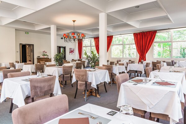Uma sala de jantar com várias mesas e cadeiras, janelas amplas com cortinas vermelhas e um candelabro.