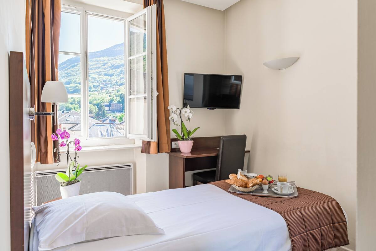 Quarto de hotel com cama, secretária com café da manhã, televisão e janela com cortinas.