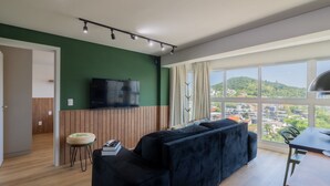 Un salon moderne avec un mur d’accent vert, un téléviseur à écran plat, un canapé foncé, une petite table ronde avec des livres et une vue sur le paysage urbain à travers de grandes fenêtres.