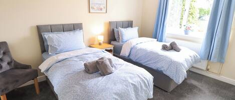 A hotel room with two beds, a nightstand, a chair, and a window with curtains.