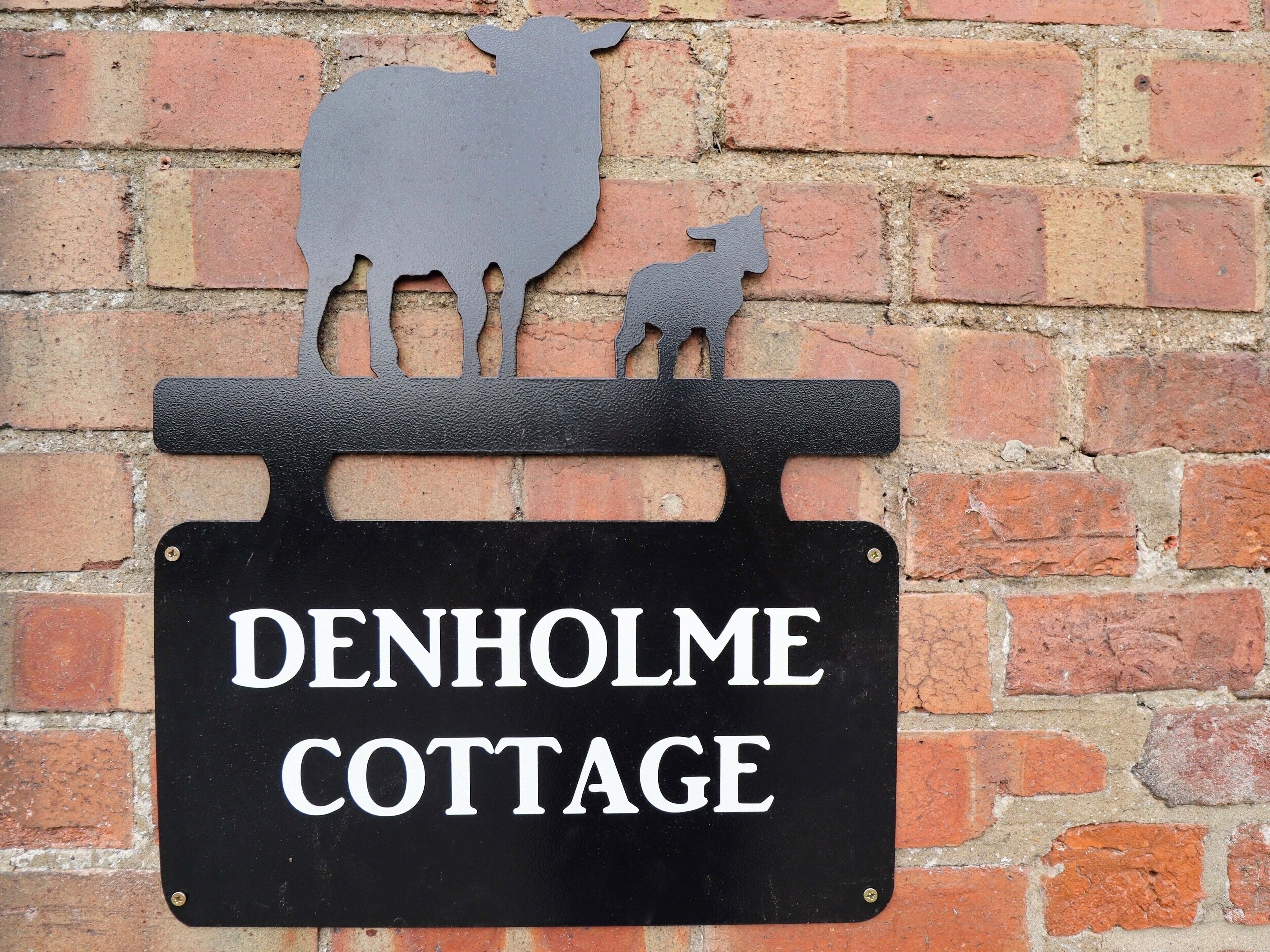 A black metal sign with white lettering for 'DENHOLME COTTAGE' mounted on a brick wall.