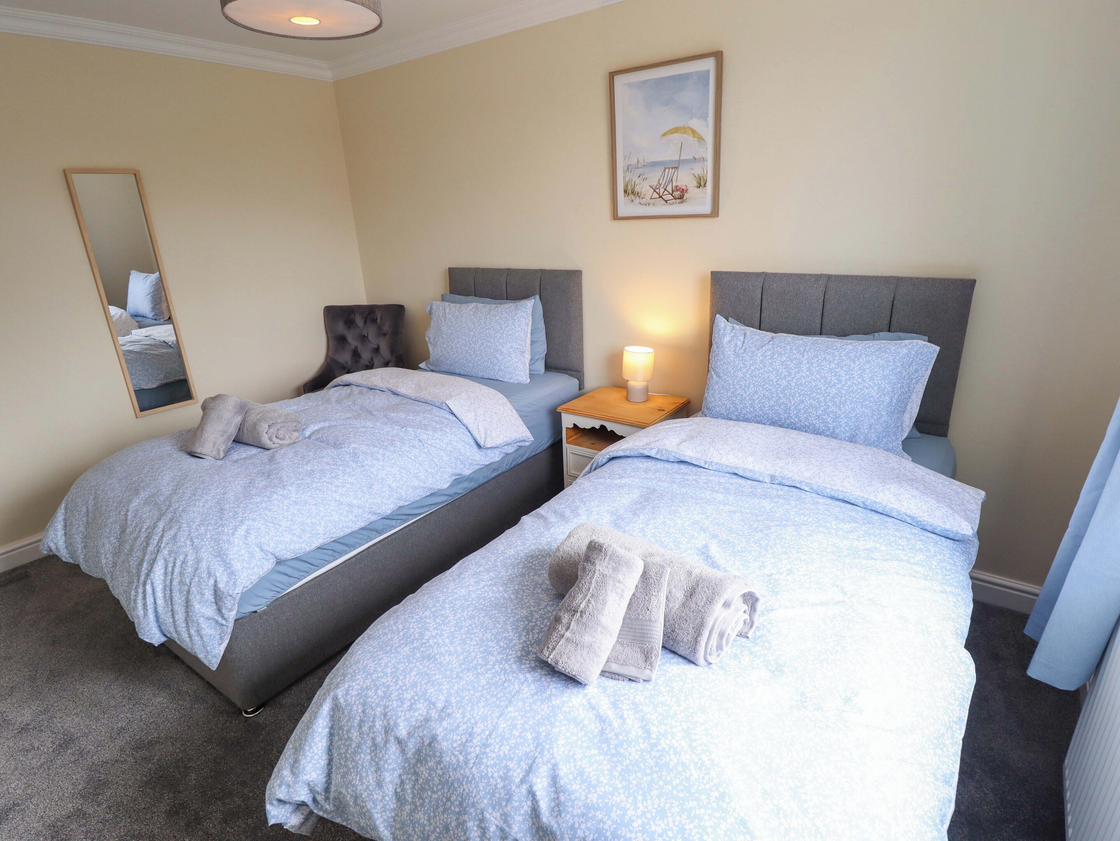 Une chambre avec deux lits jumeaux, une table de chevet avec une lampe, un miroir et un tableau sur le thème de la plage au mur.