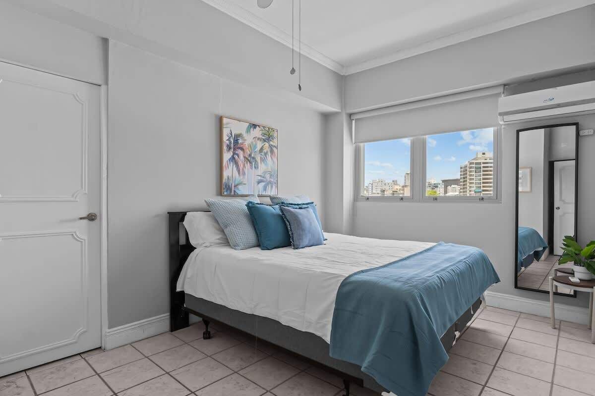 A bedroom with a bed, a window with a city view, a door, and a painting on the wall.