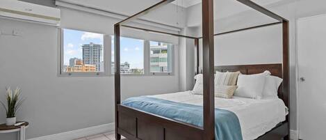 Una cama con dosel en una habitación con vistas a la ciudad, una mesita con una planta y una puerta blanca.