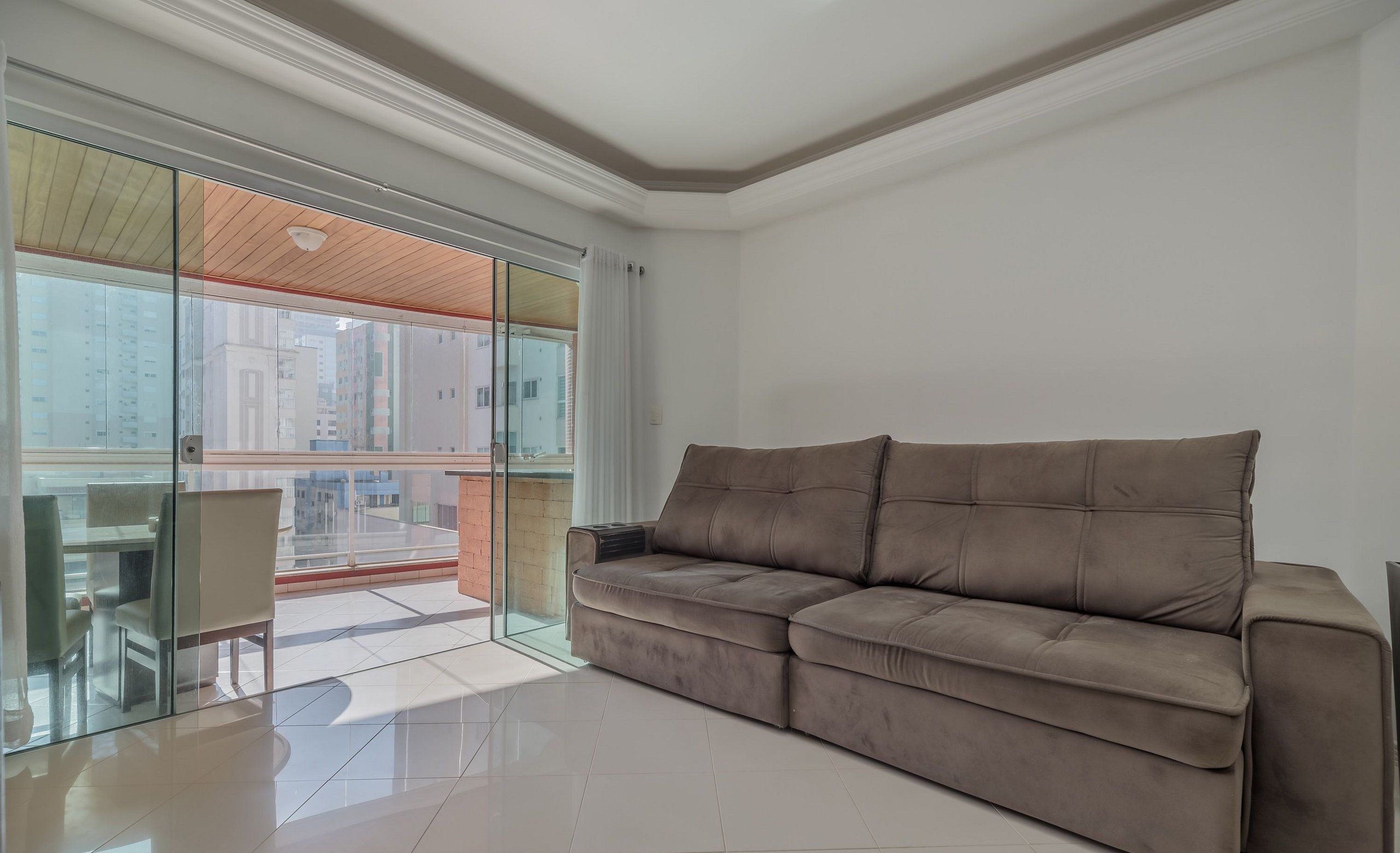 Una sala de estar moderna con un sofá marrón, un ventanal de vidrio que da a un balcón exterior y vistas de edificios al fondo.